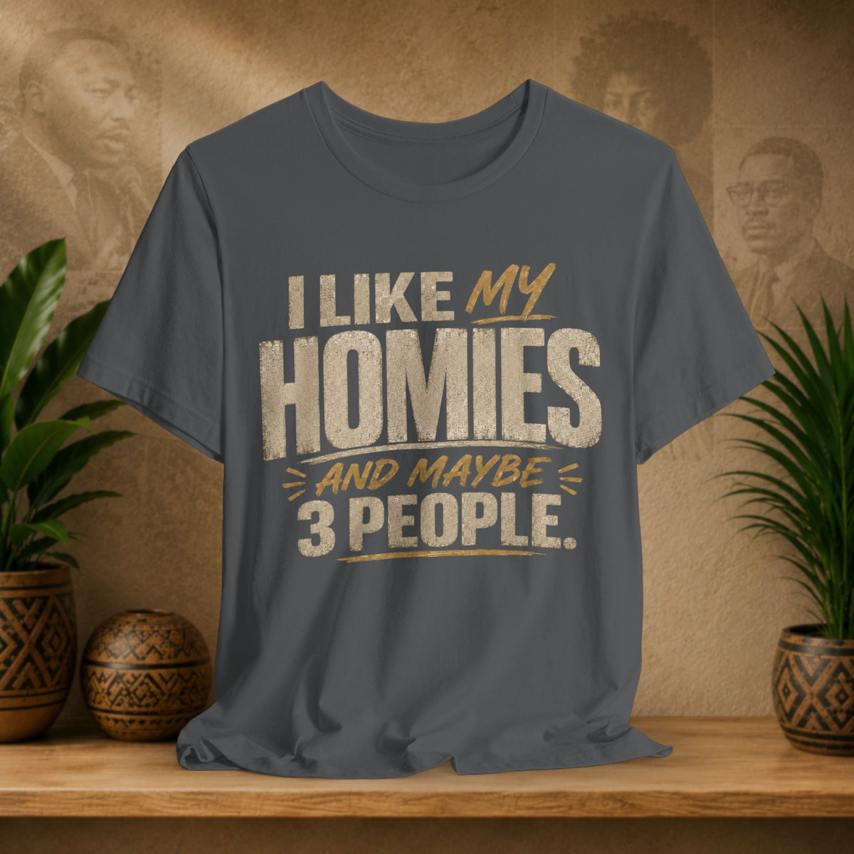 A dark grey graphic t-shirt displayed on a wooden shelf. The shirt features a humorous slogan printed in white and tan lettering: 'I LIKE MY HOMIES and maybe 3 PEOPLE.