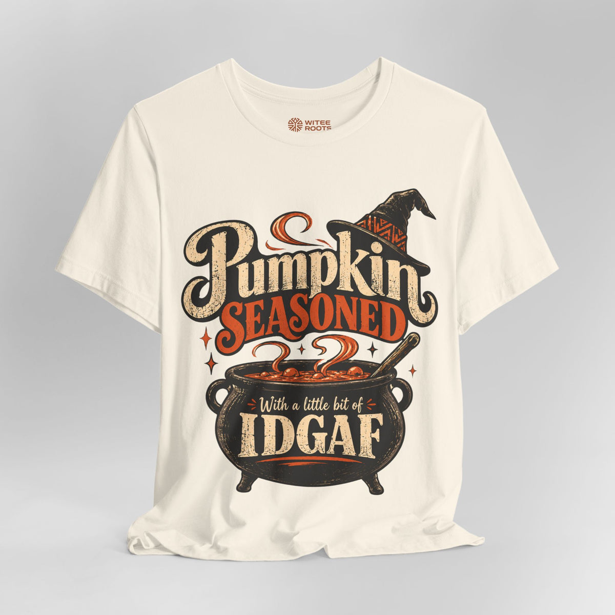 An off-white crewneck t-shirt with a Halloween graphic that reads 'Pumpkin Seasoned' and 'With a little bit of IDGAF'. The graphic features a witch's hat and a cauldron.