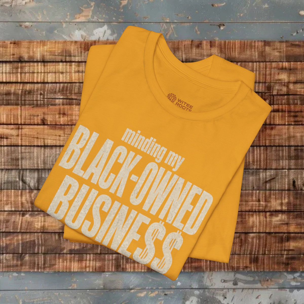 A folded mustard yellow t-shirt with a white graphic text reading 'minding my BLACK-OWNED BUSINESS' is displayed on a rustic wooden plank surface.