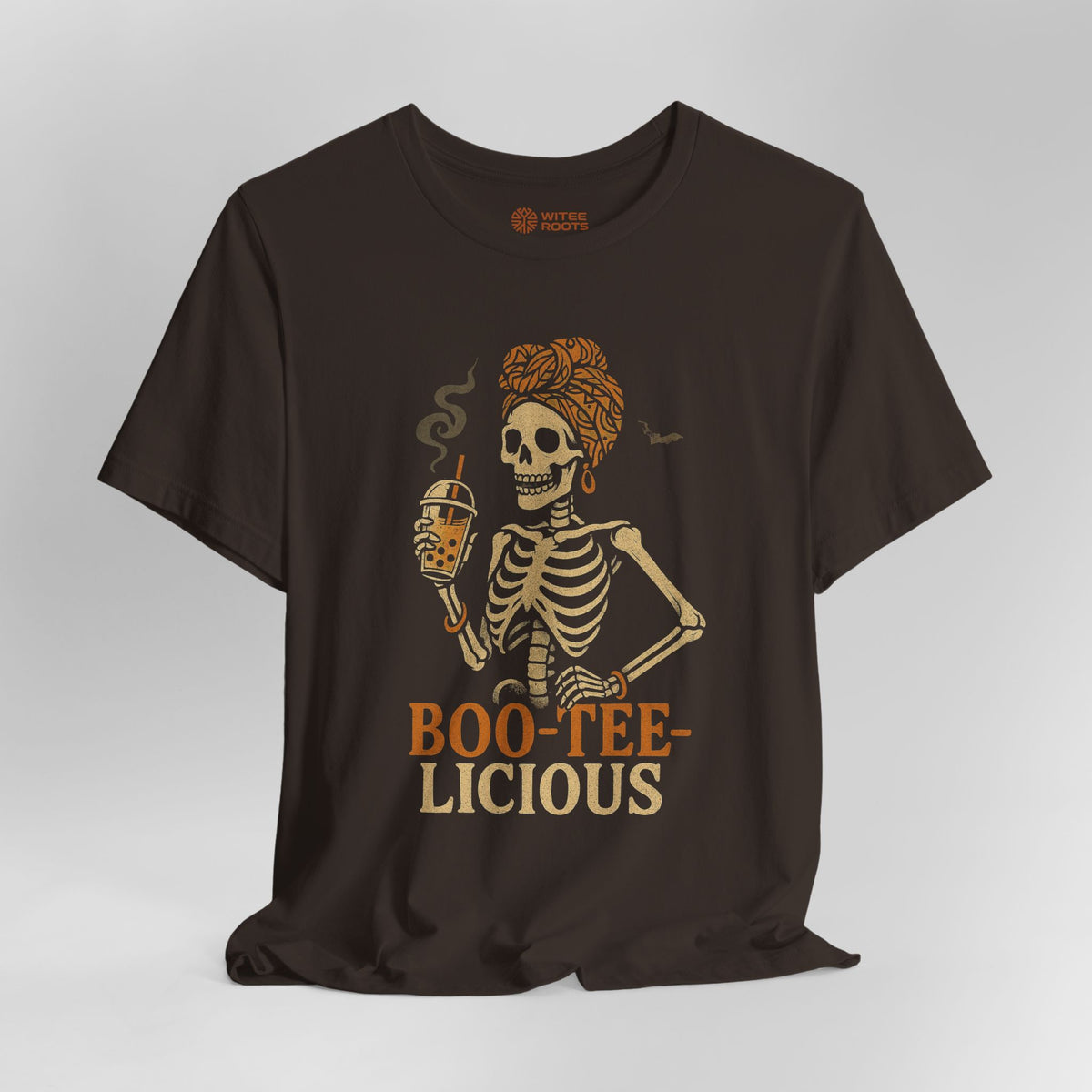 A dark brown short-sleeve t-shirt with a graphic of a skeleton woman holding a beer mug, with the text 'BOO-TEE-LICIOUS' printed below in orange and beige. The shirt is displayed against a neutral gray background.