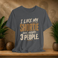 A dark grey graphic t-shirt displayed on a wooden shelf. The shirt features a humorous slogan printed in white and tan lettering: 'I LIKE MY SHORTIE and maybe 3 PEOPLE.