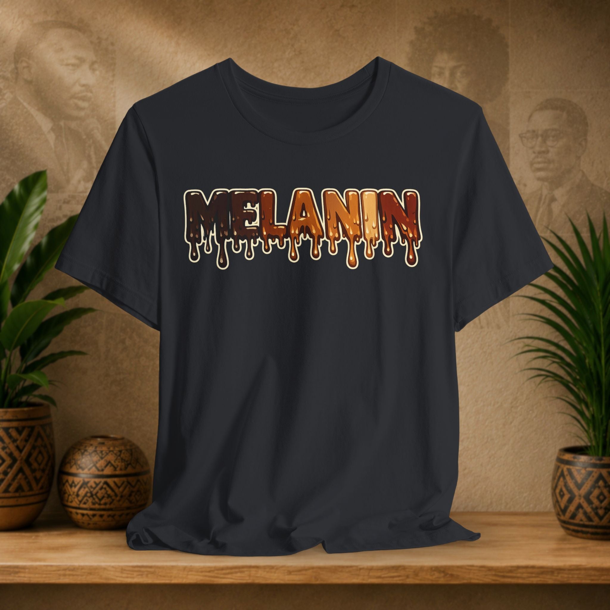 A black t-shirt with a 'MELANIN' text graphic in a dripping paint style, displayed on a wooden shelf with potted plants and a textured background.