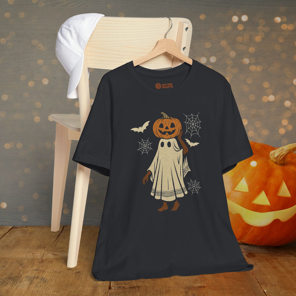 A black short-sleeve t-shirt with a cartoon pumpkin ghost Halloween graphic, hanging on a wooden hanger on a light wood chair. A carved orange pumpkin and a white cap are styled in the background against a bokeh light gray wall.
