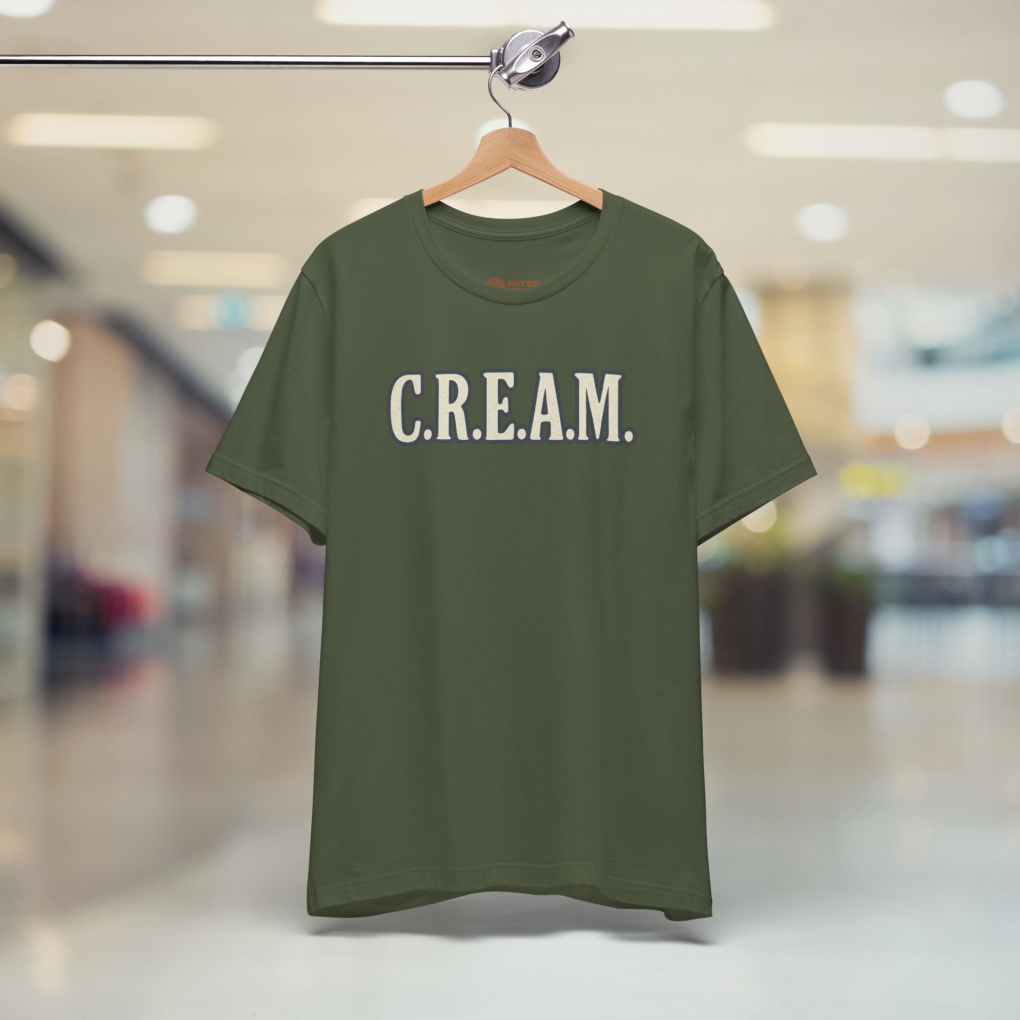 Green t-shirt with 'C.R.E.A.M.' text on a hanger in a blurred indoor setting
