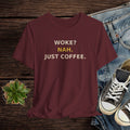 Maroon t-shirt with 'WOKE? NAH. JUST COFFEE.' text on a wooden surface with jeans and sneakers.