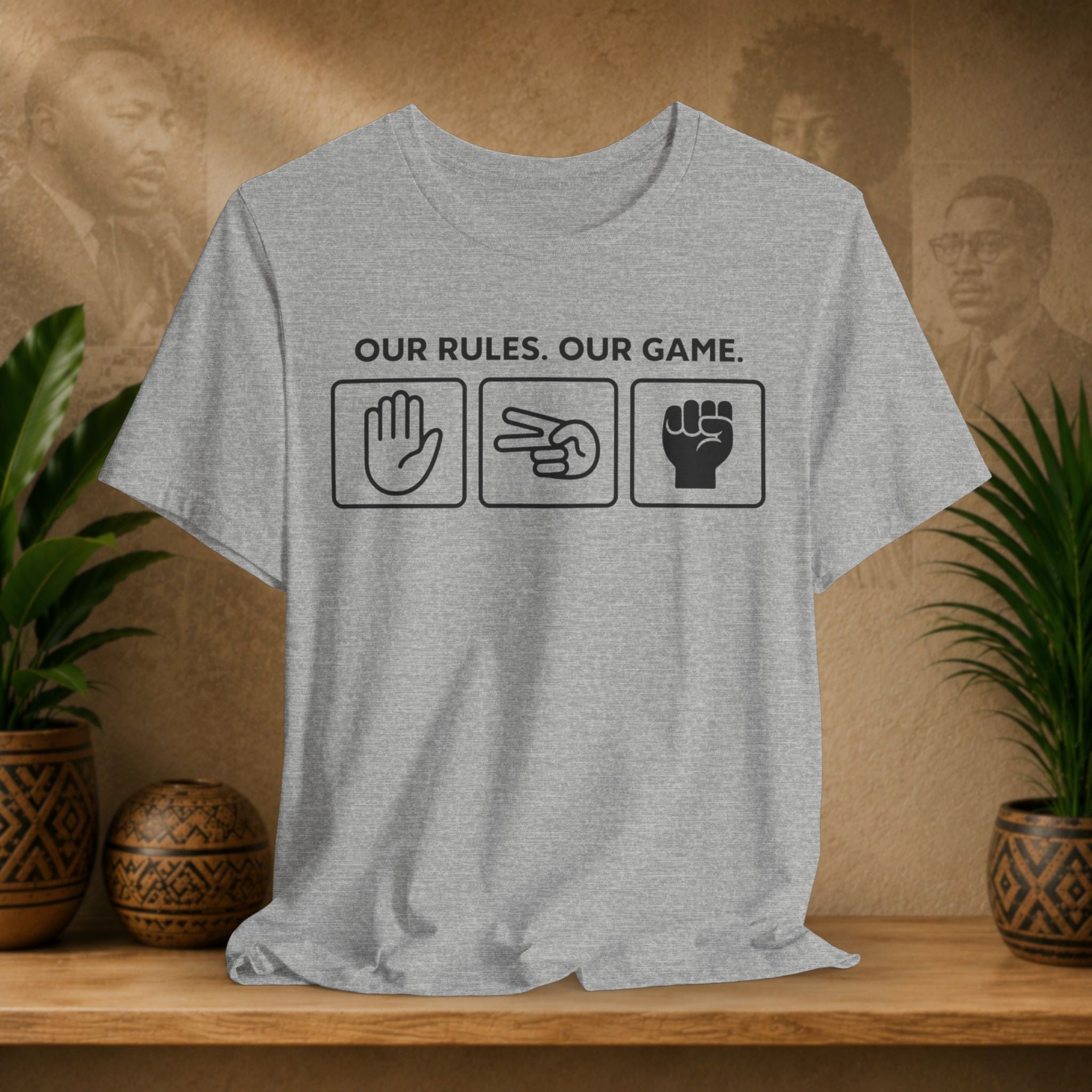 Gray t-shirt with 'Our Rules, Our Game' text and symbols on a wooden surface with plants and a decorative item in the background.