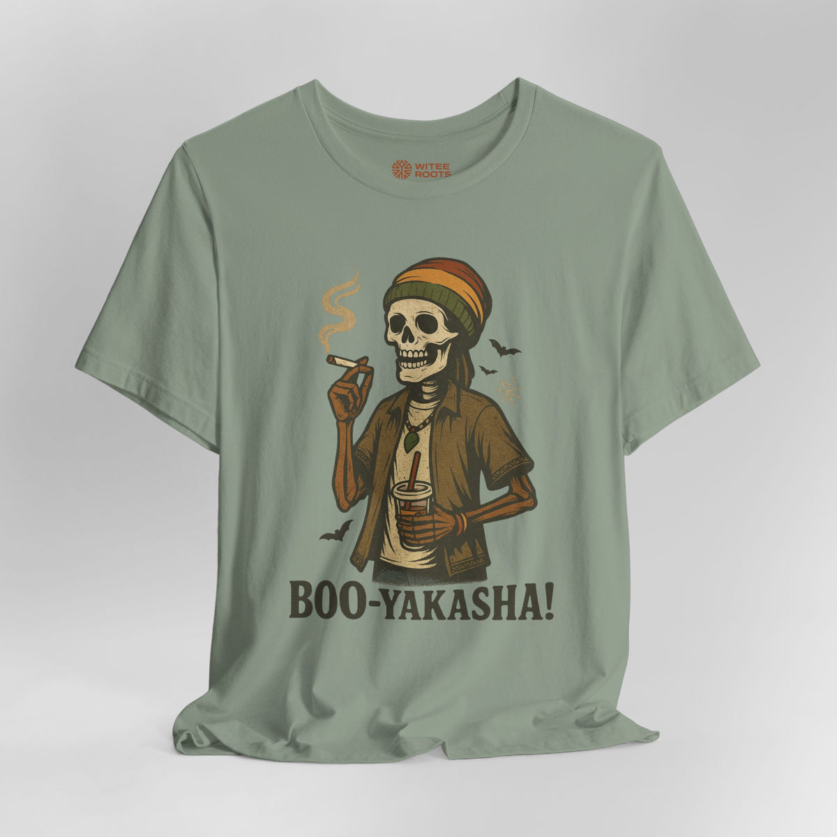 A sage green graphic t-shirt featuring a cartoon illustration of a skeleton in a Rasta hat smoking and holding a drink, with the text 'BOO-YAKASHA!' printed below. The t-shirt is displayed against a neutral light gray background.