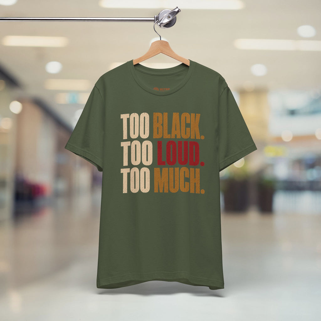 An olive green graphic t-shirt hanging on a wooden hanger from a clothing rack, set against a blurred indoor hallway background. The shirt features the text 'TOO BLACK.
