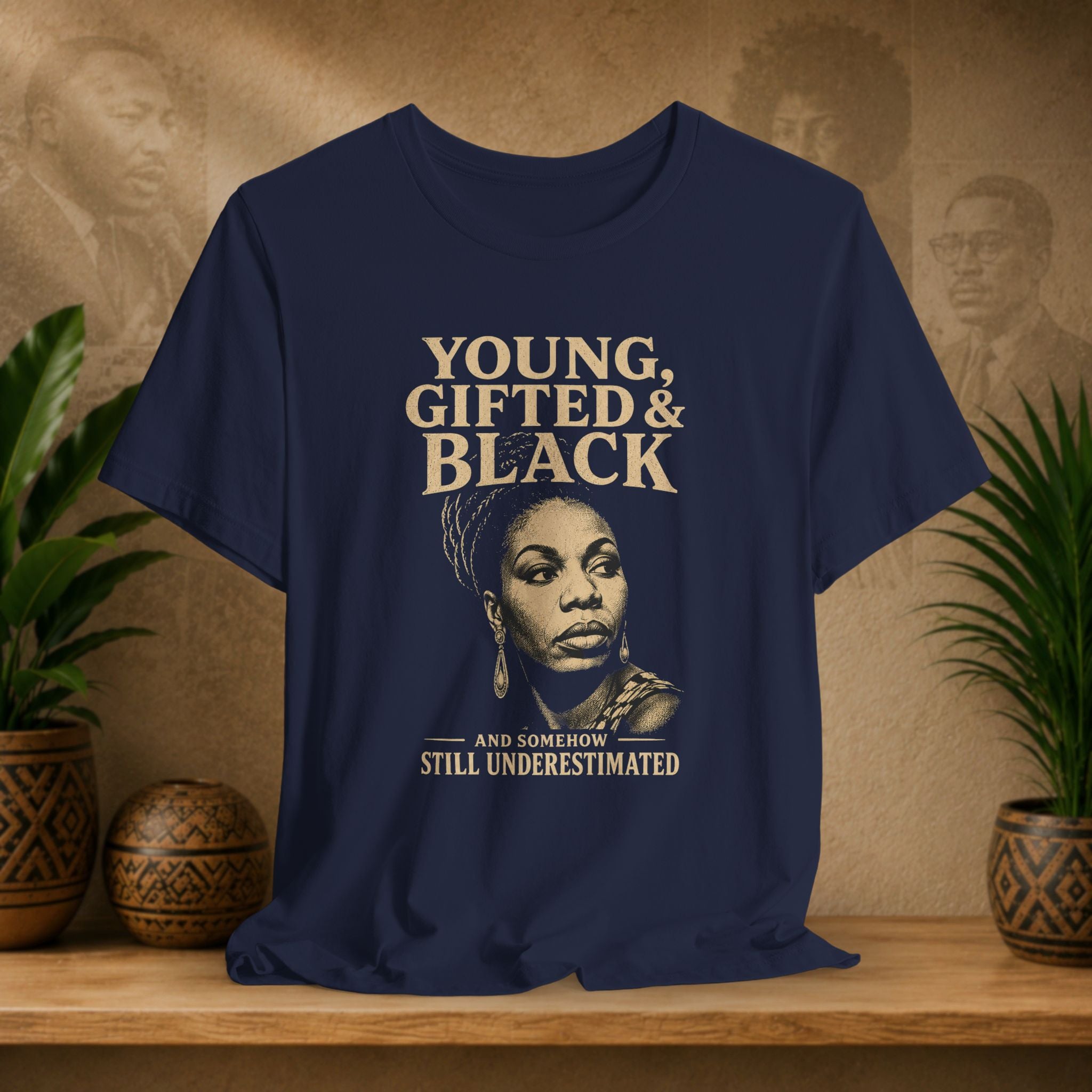 A navy blue graphic t-shirt displayed on a wooden shelf. The shirt features a beige and black portrait of Audie Lushan and the text 'YOUNG, GIFTED & BLACK' and 'AND SOMEBOW STILL UNDERESTIMATED'.