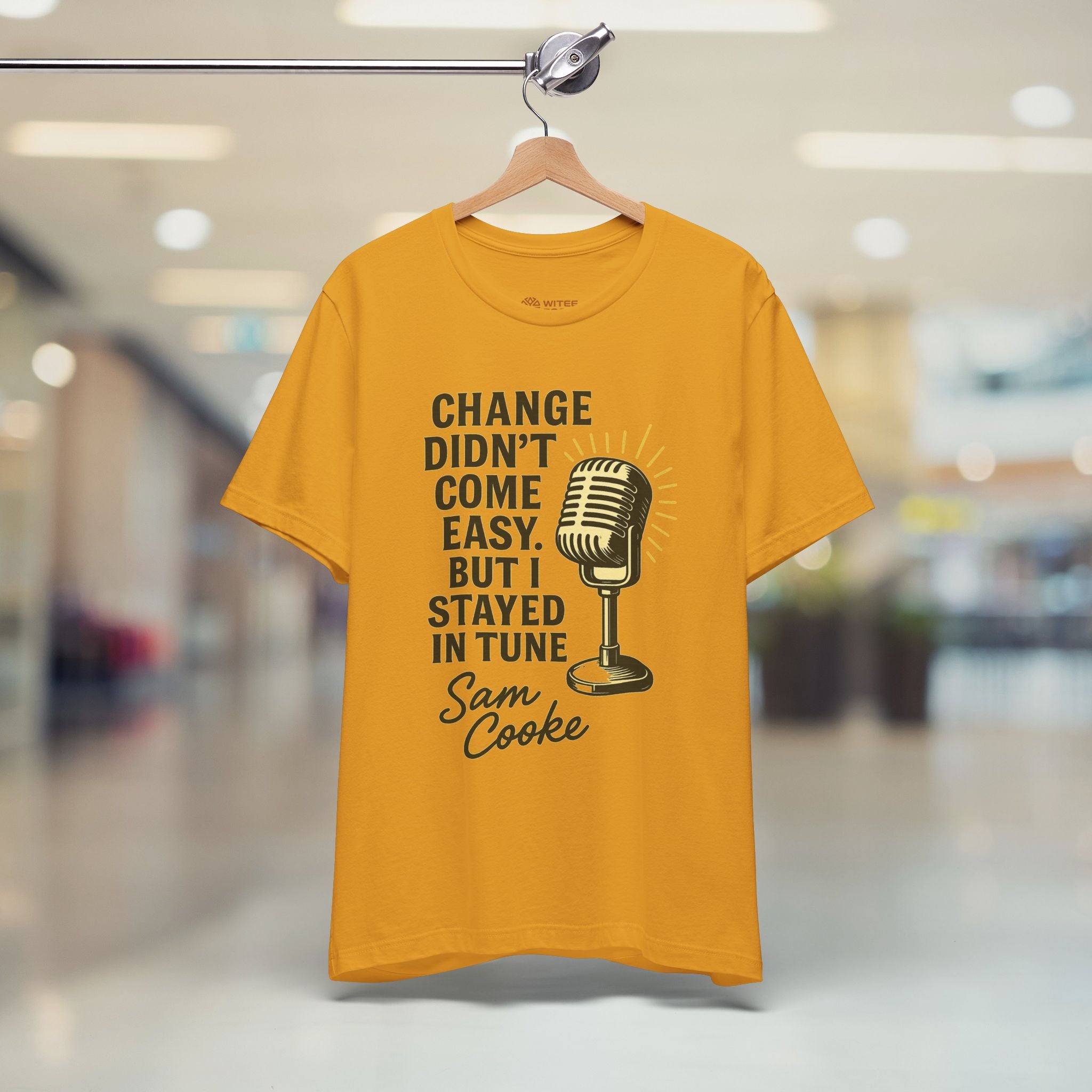 Mustard t-shirt with a microphone graphic and text on a hanger in a store.
