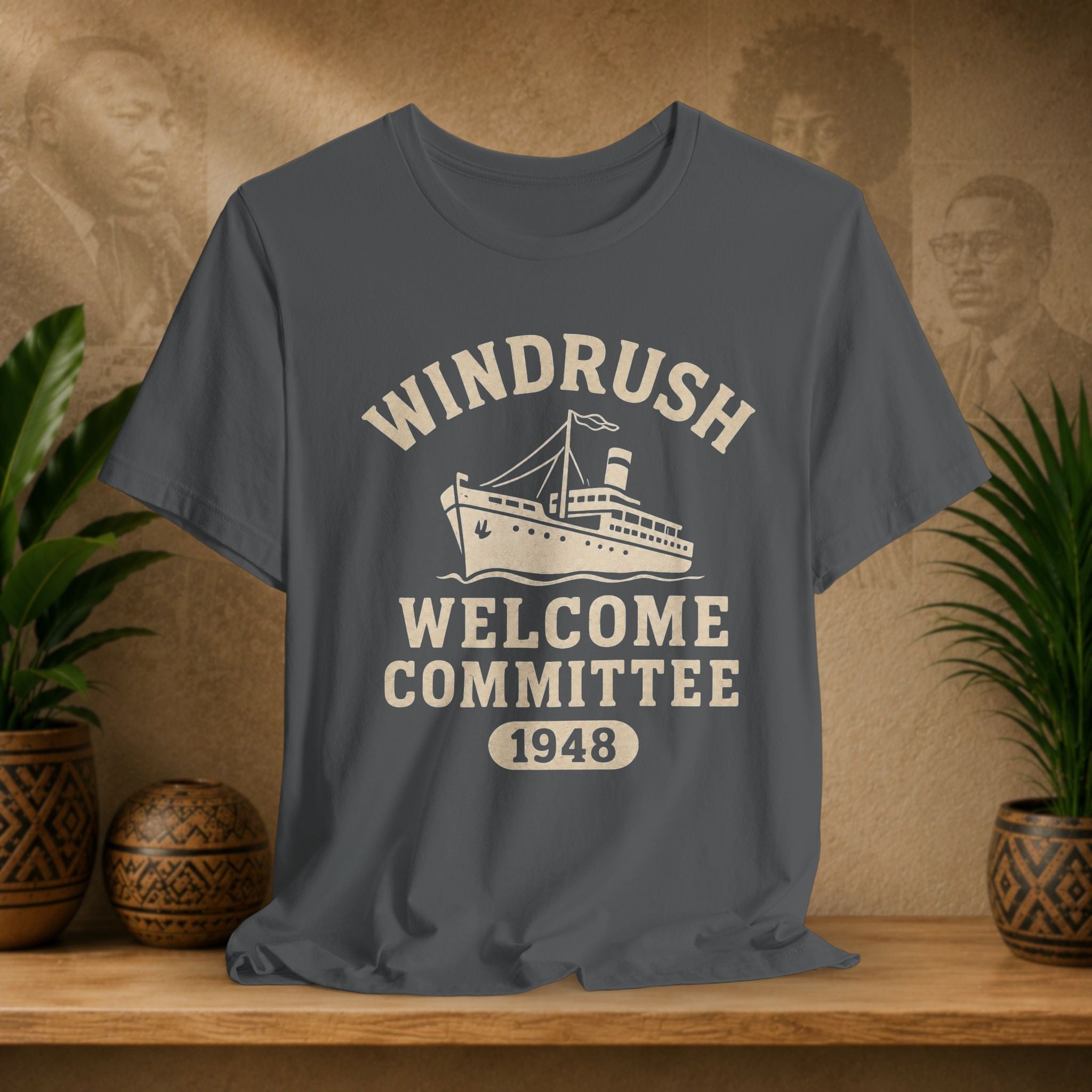 Gray t-shirt with 'Windrush Welcome Committee 1948' design on a wooden surface with plants and a decorative wall.