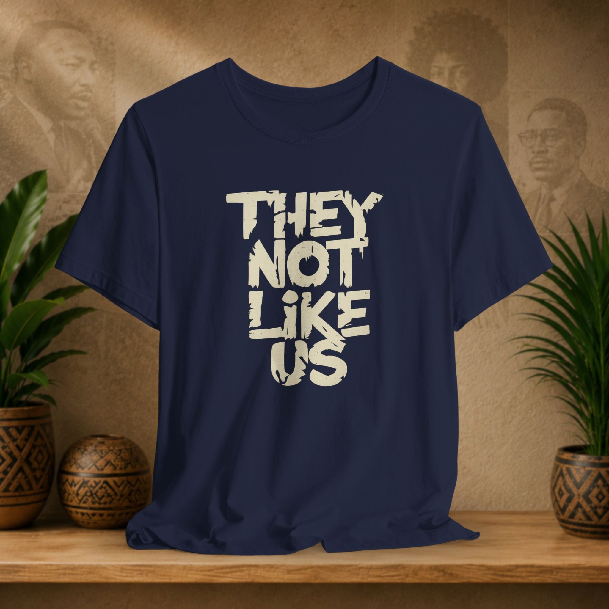 Navy blue t-shirt with 'THEY NOT LIKE US' text on a wooden surface with plants and a textured wall background.