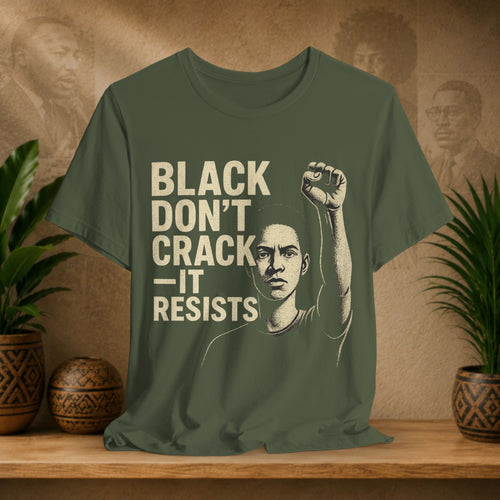 Green t-shirt with a graphic and text 'Black Don't Crack, It Resists' on a wooden surface with a decorative background.