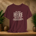 Maroon t-shirt with white text on a wooden surface with plants and a textured wall background