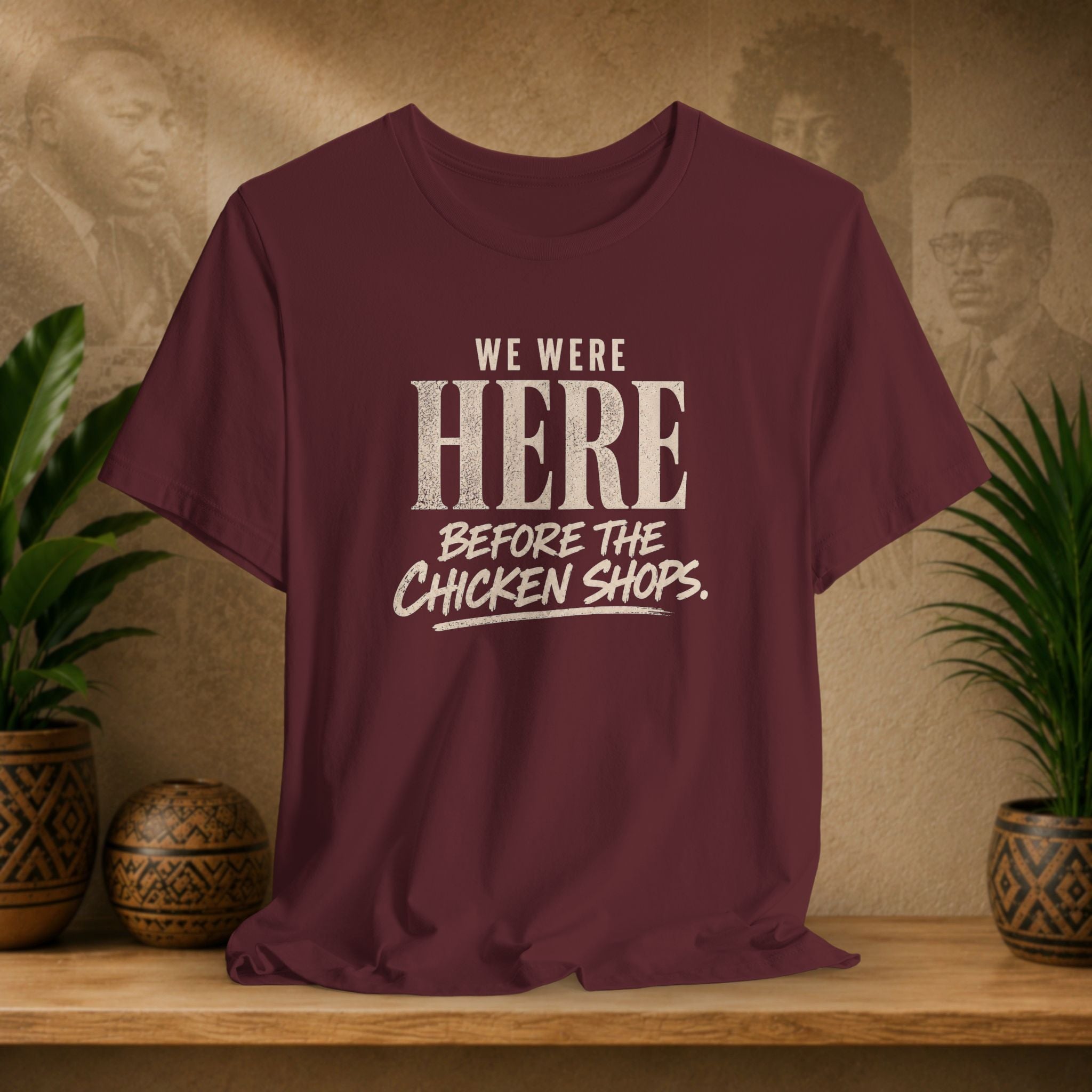 Maroon t-shirt with white text on a wooden surface with plants and a textured wall background