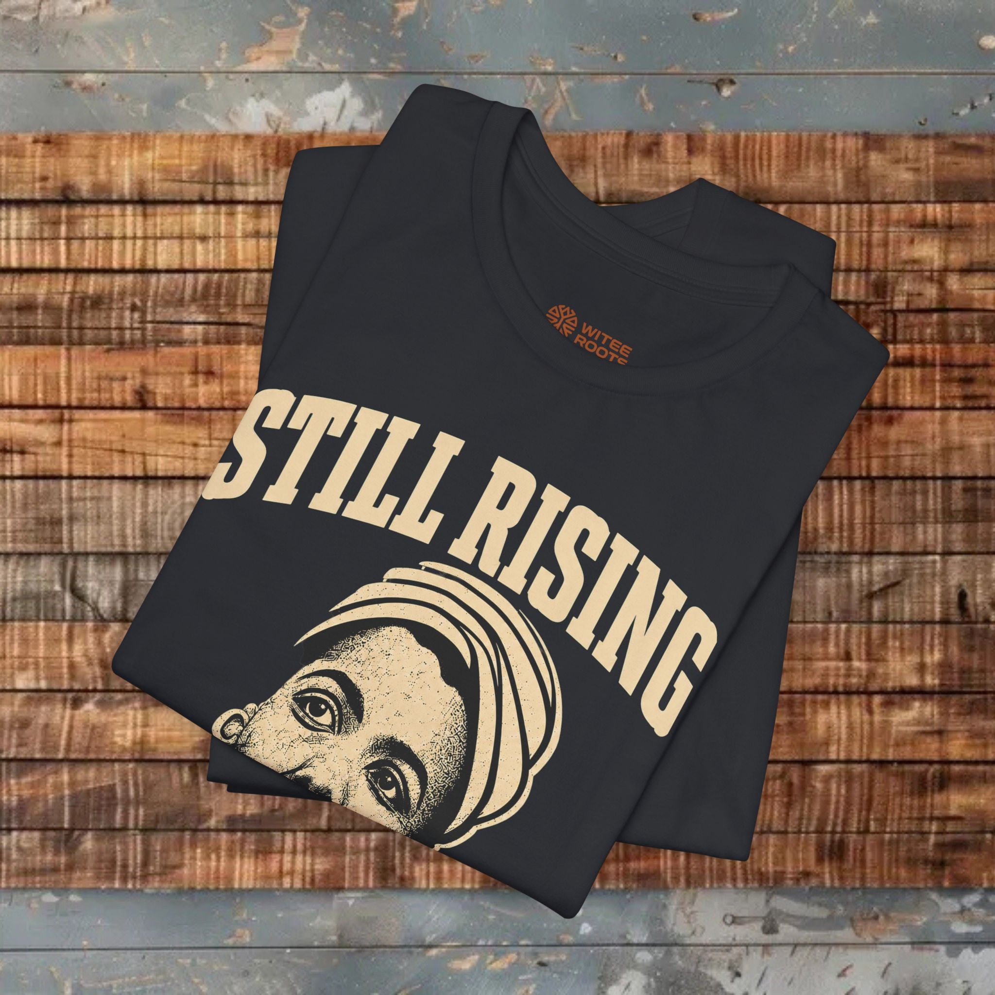 Black t-shirt with 'STILL RISING' text and graphic on a wooden surface