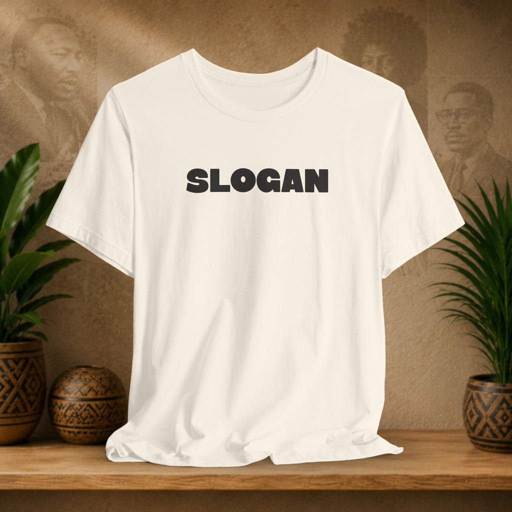 A beige short-sleeve crewneck t-shirt with the word 'SLOGAN' printed in bold black letters across the chest, displayed on a wooden shelf with potted plants in the background.