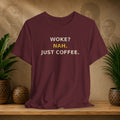 Maroon t-shirt with text 'WOKE? NAH. JUST COFFEE.' on a wooden surface with plants and wall art in the background.
