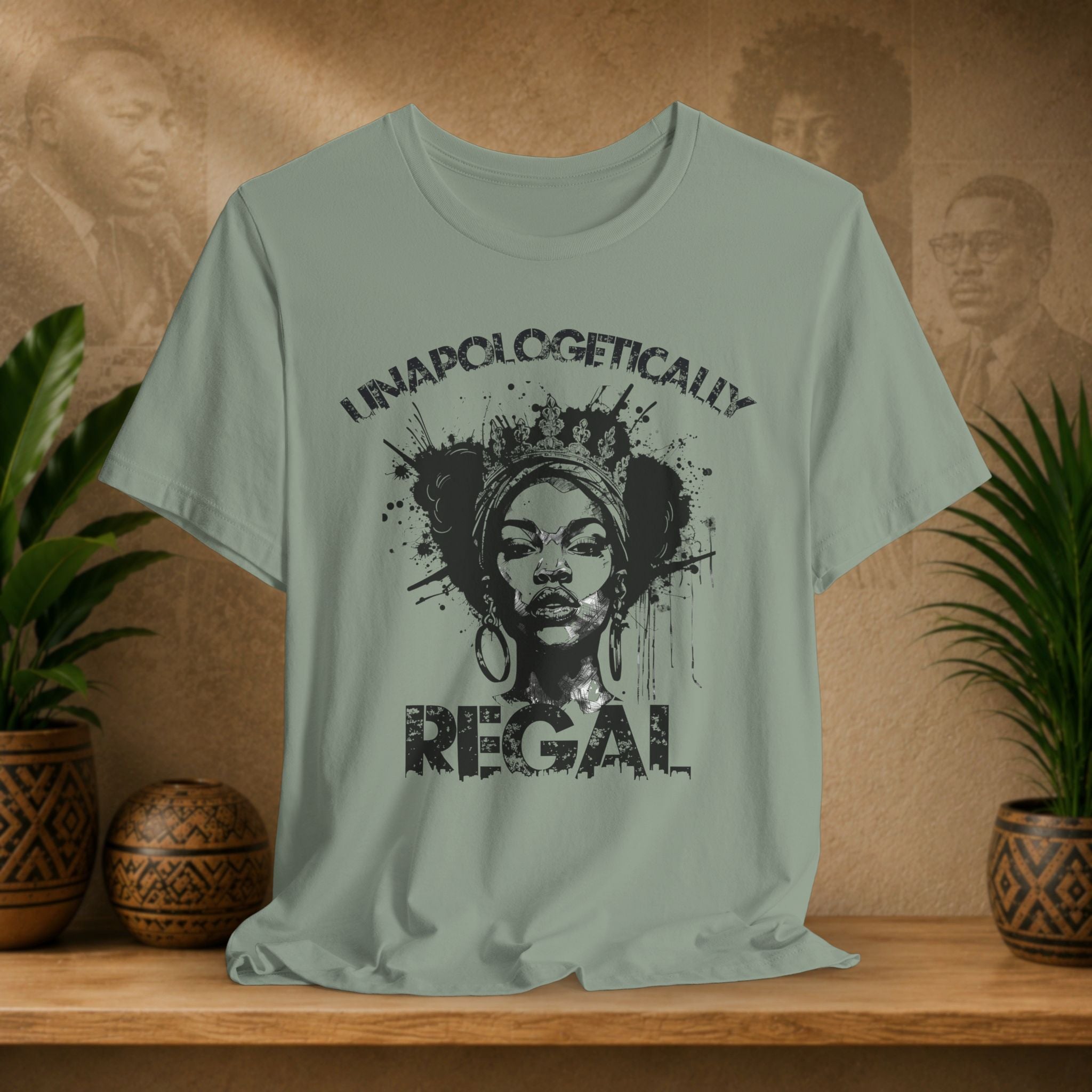 A sage green graphic t-shirt displayed on a wooden shelf, flanked by potted plants. The shirt features a black distressed graphic of a woman's face with a crown and the text 'UNAPOLOGETICALLY REGAL'.