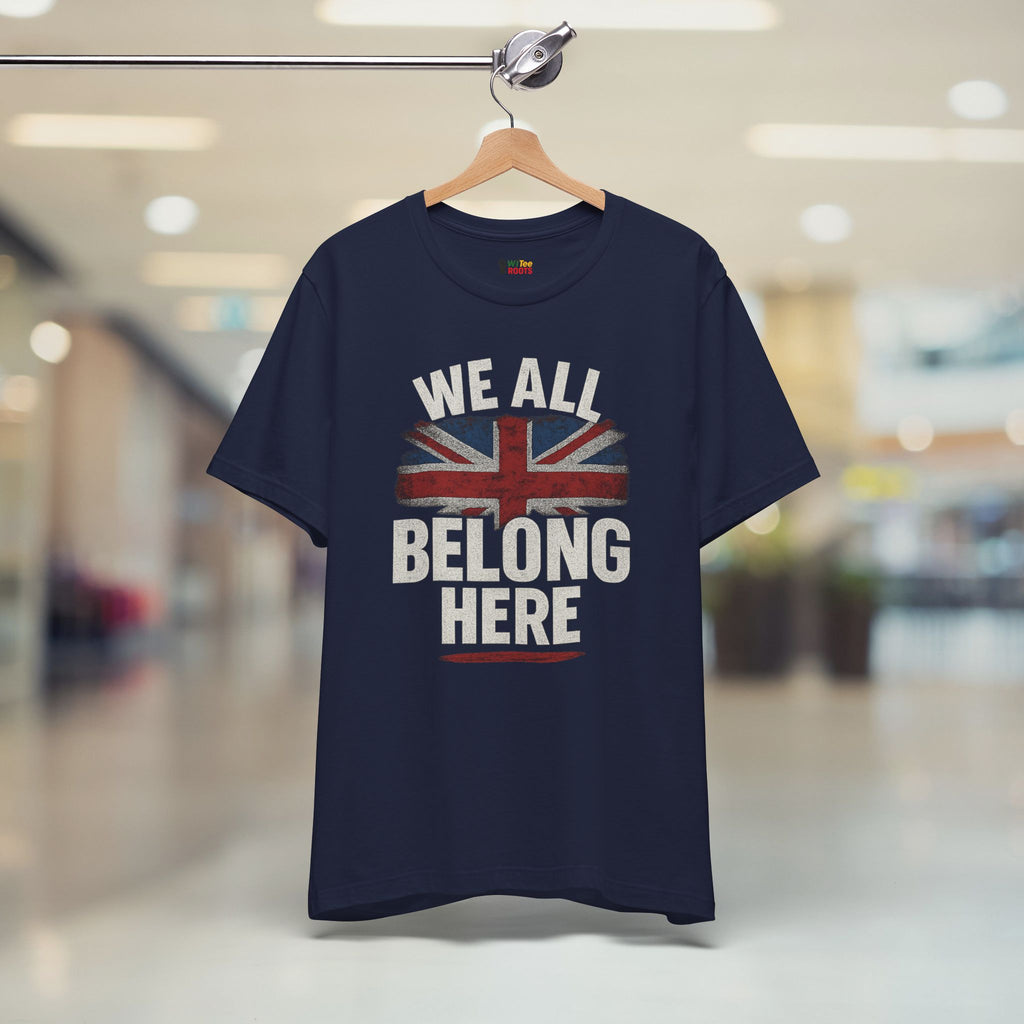 Navy blue t-shirt with Union Jack design and 'We All Belong Here' text on a hanger in a blurred indoor setting.
