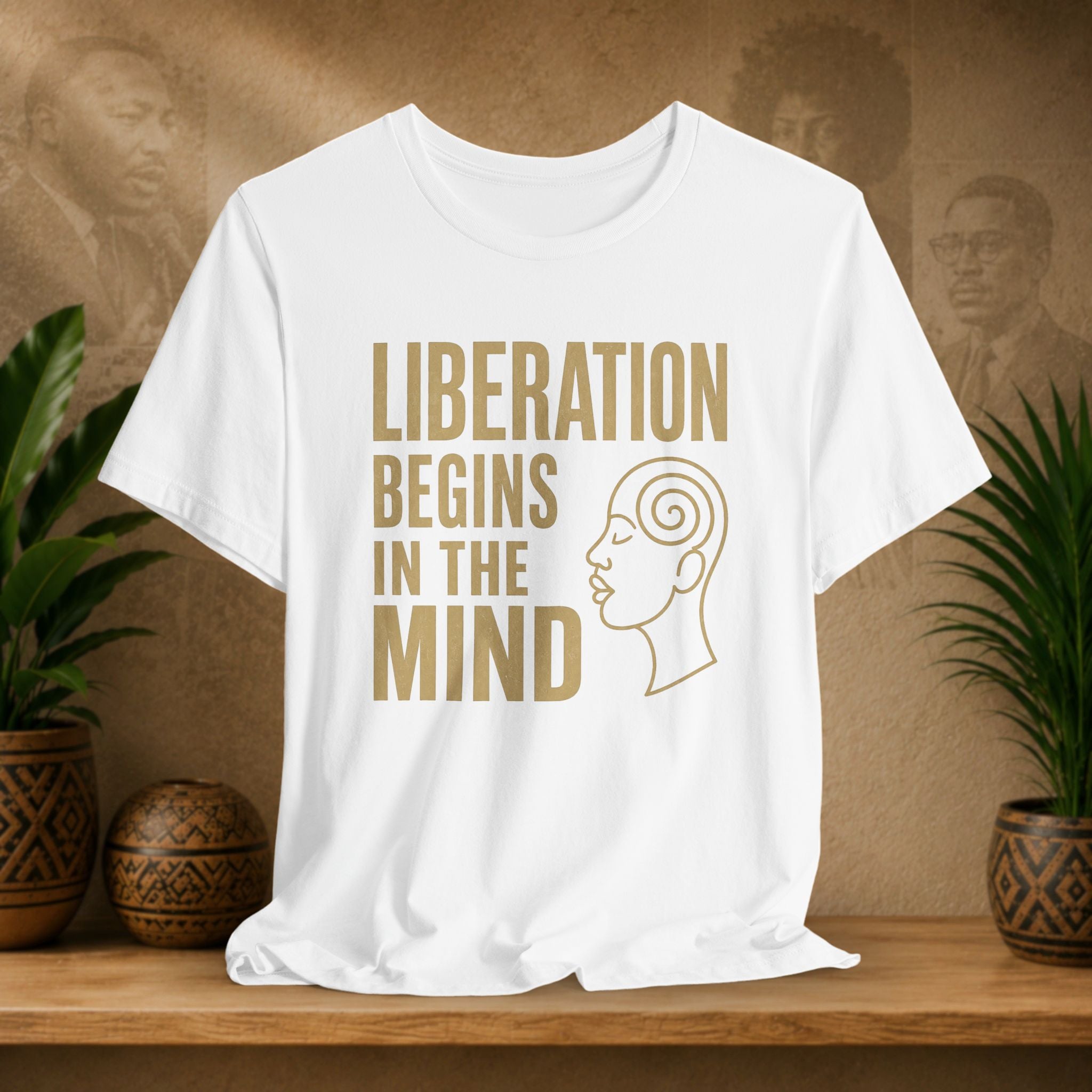 White t-shirt with 'Liberation Begins in the Mind' text and a face outline on a wooden surface with plants and a textured wall background.