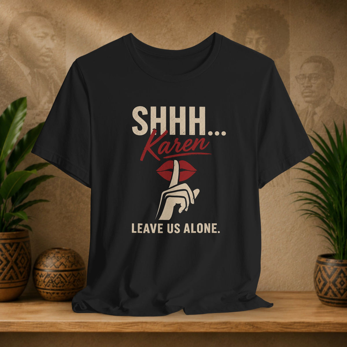 A black graphic t-shirt displayed on a wooden shelf, flanked by potted green plants against a textured beige background with vintage portraits. The shirt features a graphic print that reads 'SHHH.