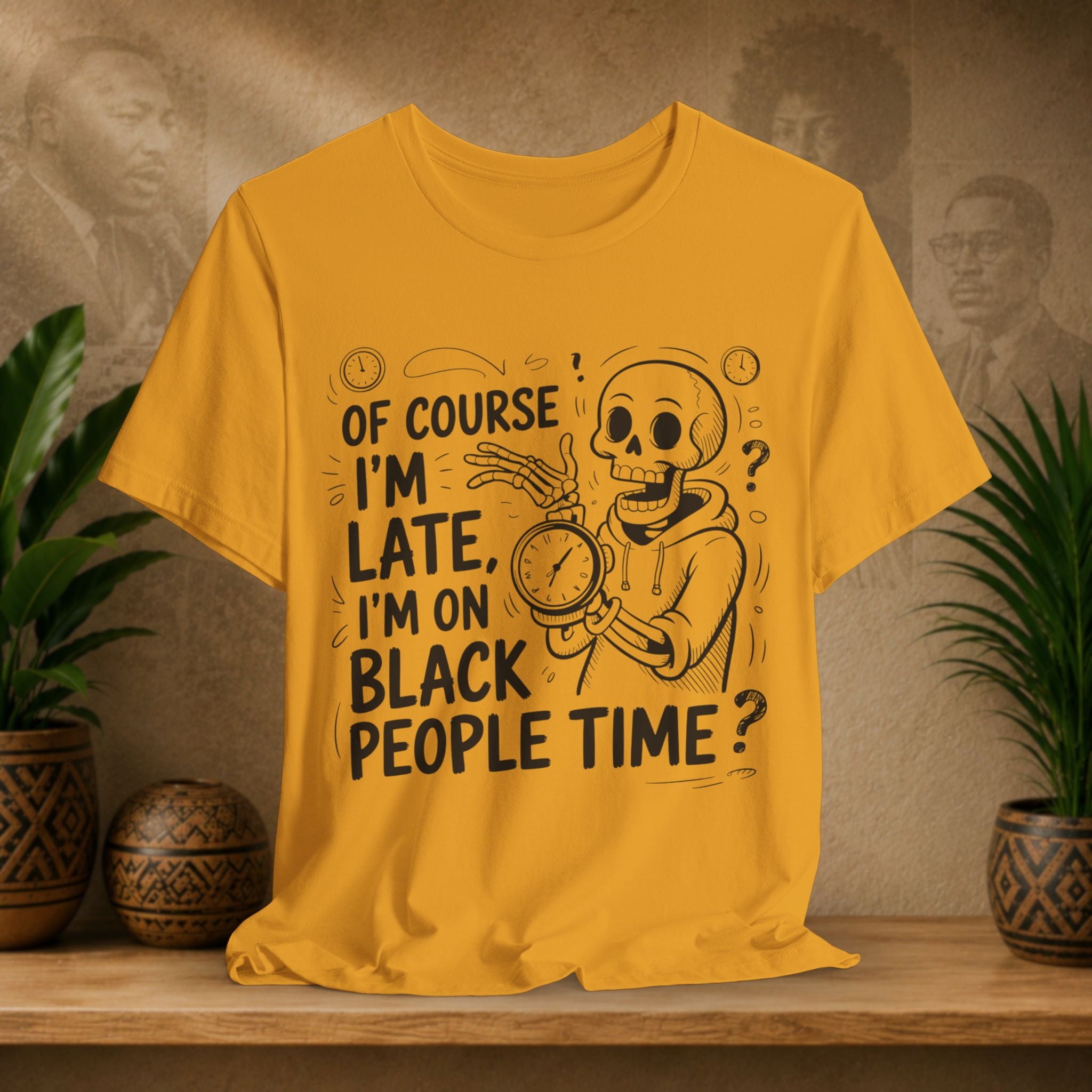 A mustard graphic t-shirt displayed on a wooden shelf, flanked by potted plants. The shirt features a black cartoon illustration of a skeleton in a hoodie holding a clock, with the text 'OF COURSE I'M LATE, I'M ON BLACK PEOPLE TIME?'.