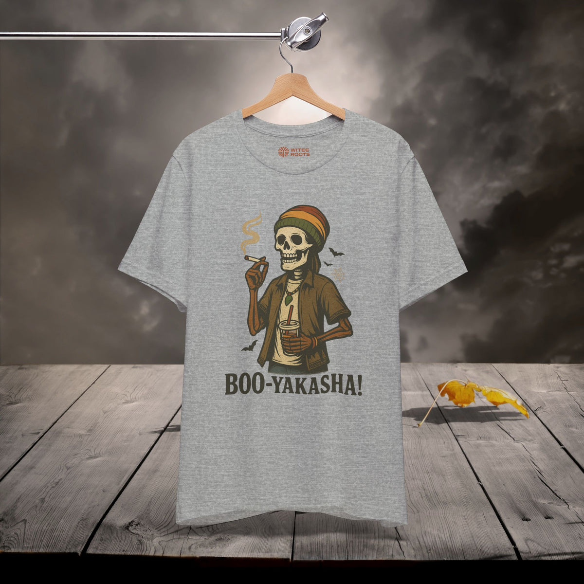 A heather gray graphic t-shirt hanging on a wooden hanger against a dark, smoky background with a rustic wooden plank floor. The graphic features a cartoon skeleton wearing a Rastafarian hat and holding a cigarette, with the text 'BOO-YAKASHA!' printed below.