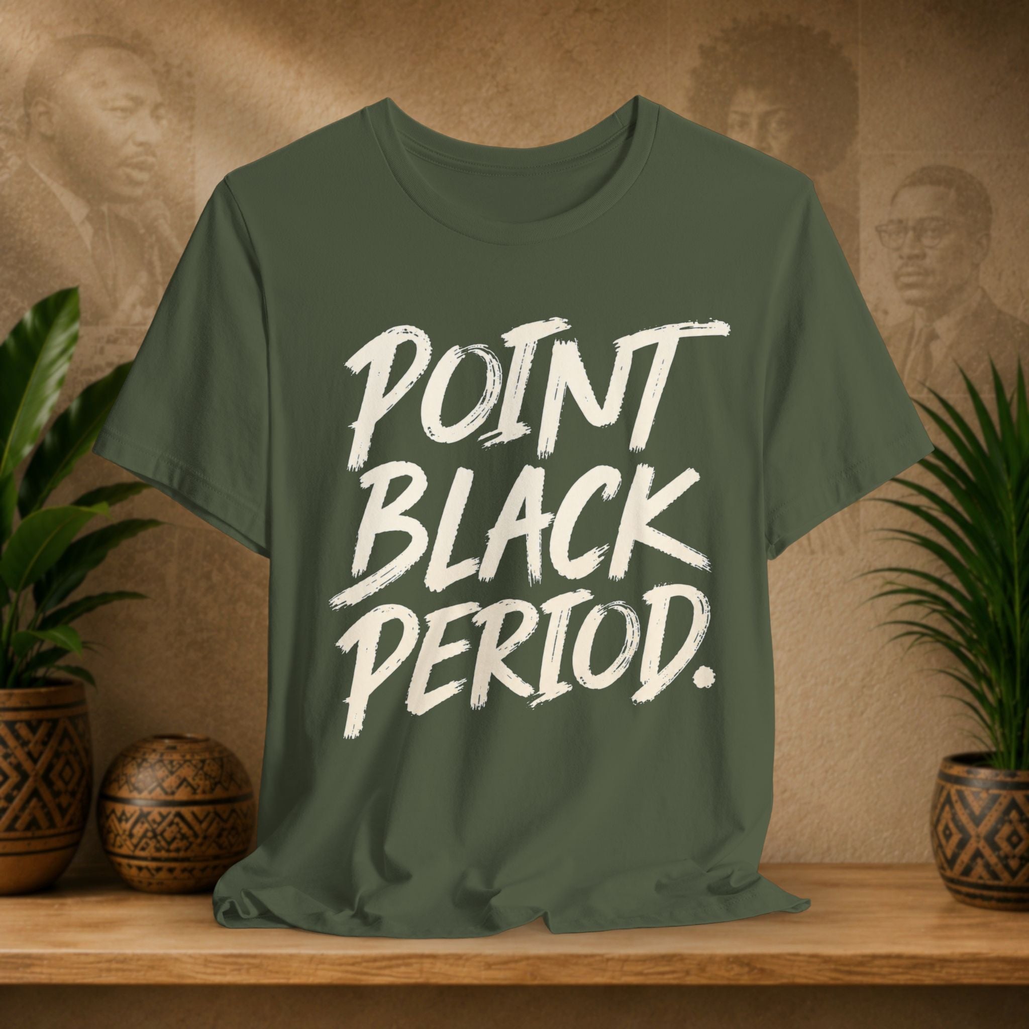 An olive green t-shirt with the text 'POINT BLACK PERIOD.' printed in a white brush-stroke font, displayed on a wooden shelf with potted plants and a textured background.