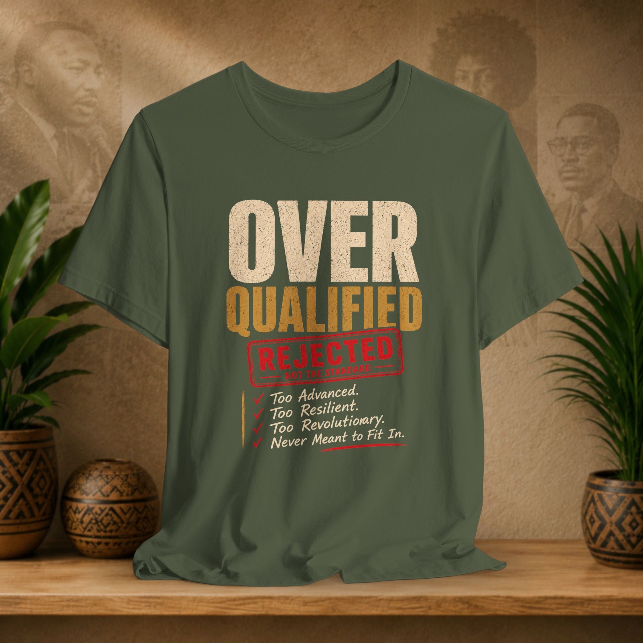 Green t-shirt with 'Over Qualified' text on a wooden surface with plants and a textured wall background