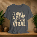 Gray t-shirt with 'I have a meme and it went viral' text on a wooden surface with plants and decorative items.