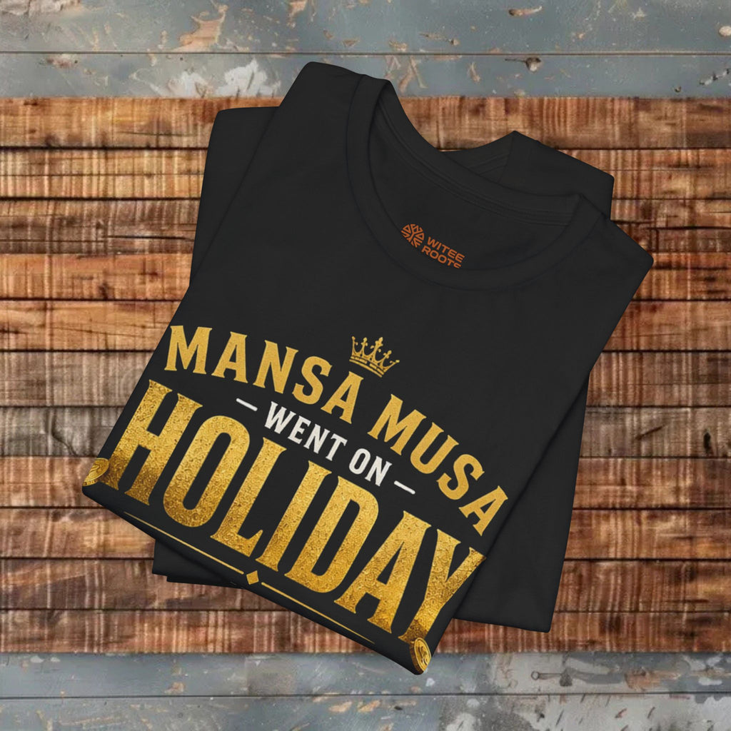 Black t-shirt with gold text 'Mansa Musa Went on Holiday' on a wooden surface