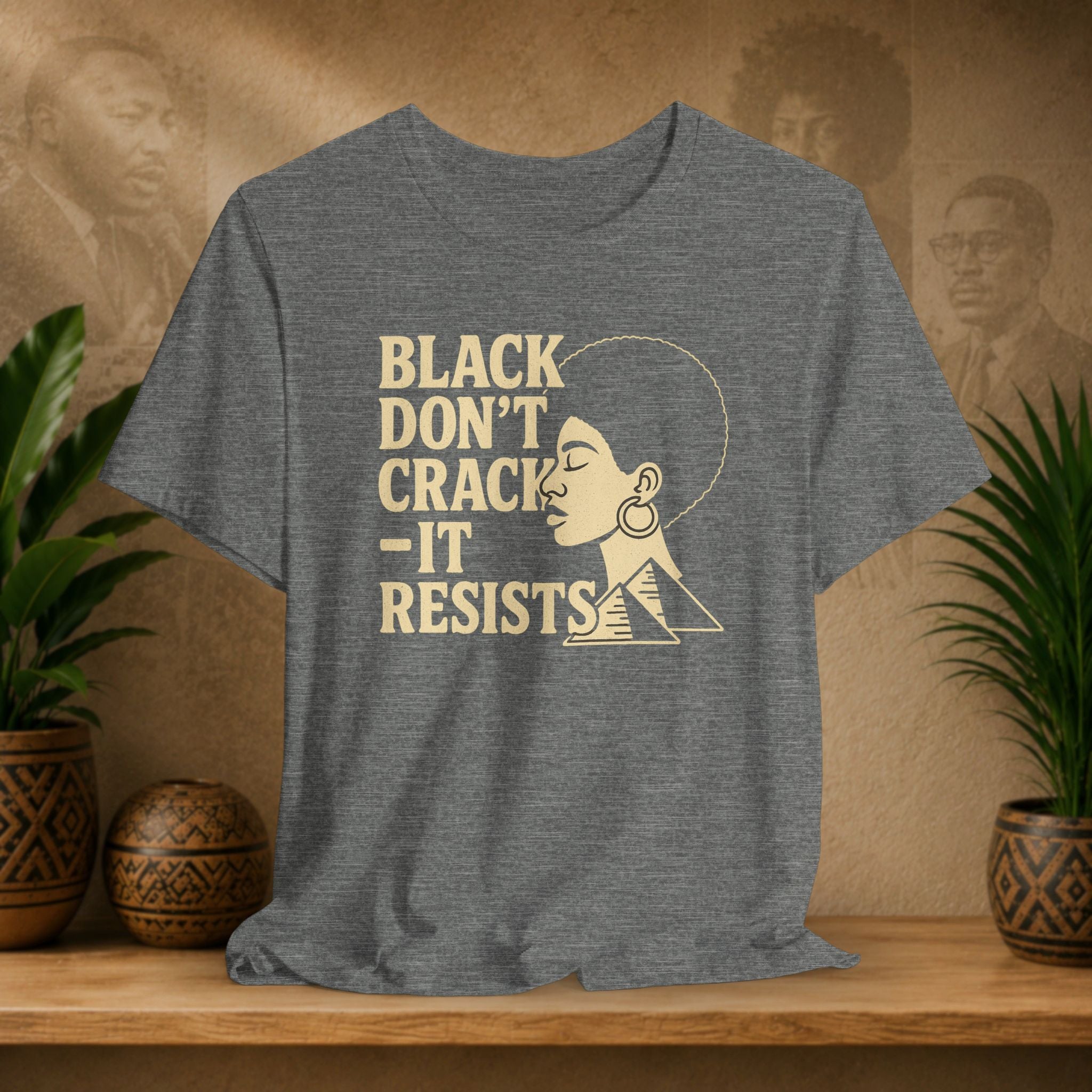 Gray t-shirt with 'Black Don't Crack, It Resists' text and graphic on a wooden surface with plants and decorative items.