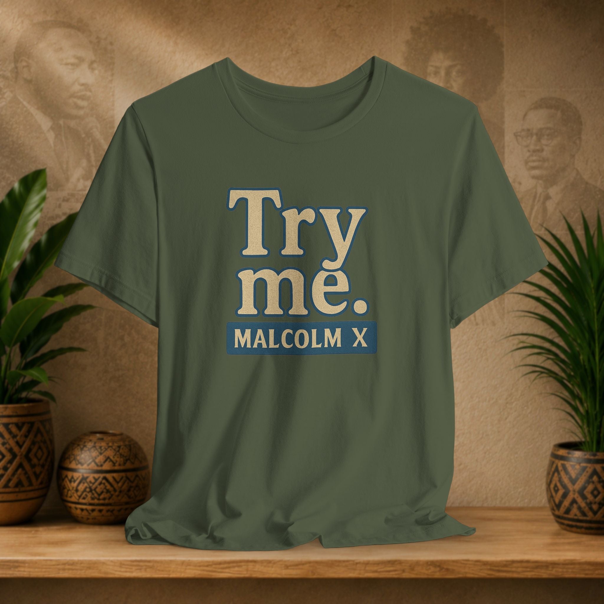 Green t-shirt with 'Try me. MALCOLM X' text on a wooden surface with decorative plants and background.