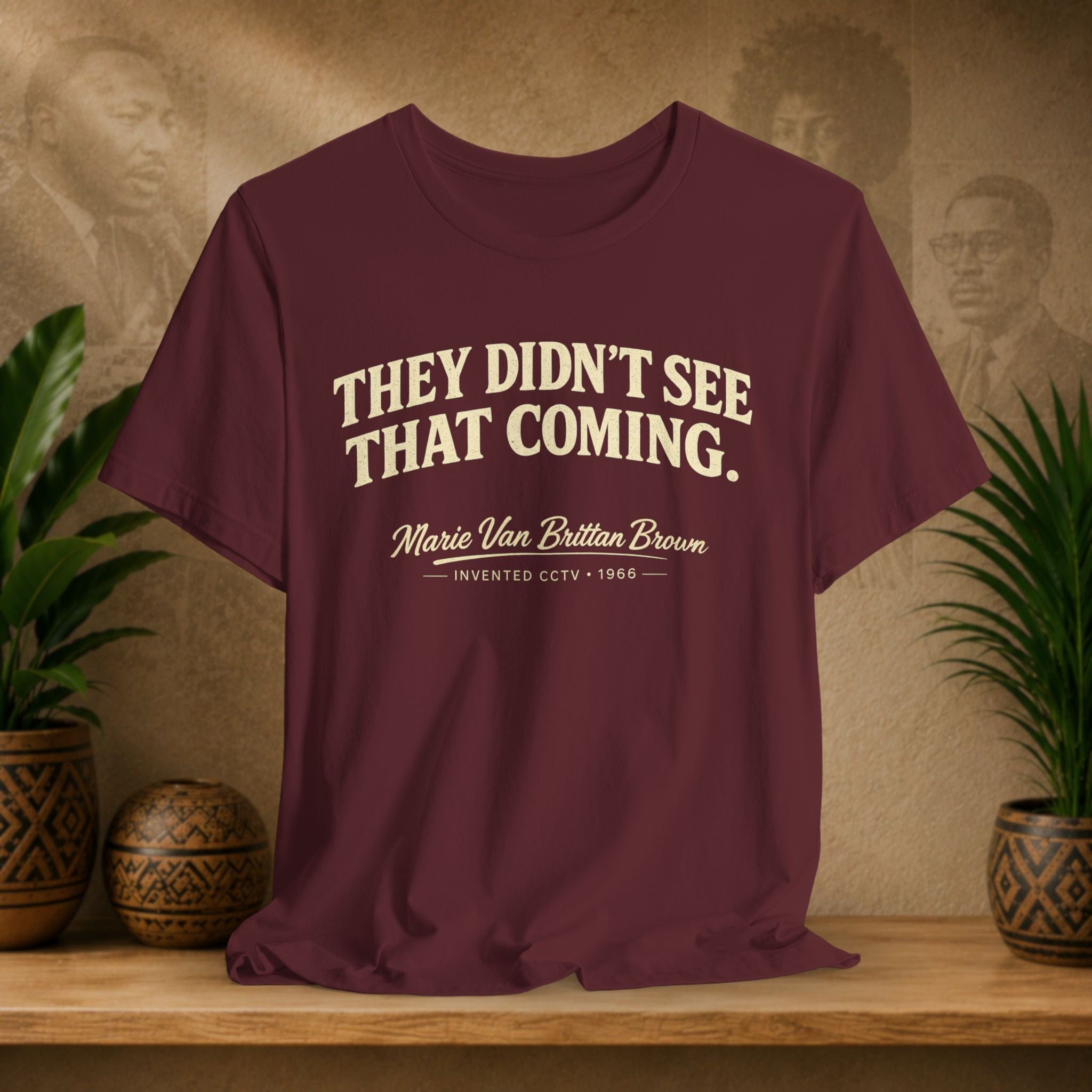 Maroon t-shirt with text on a wooden surface with plants and a wall with portraits in the background