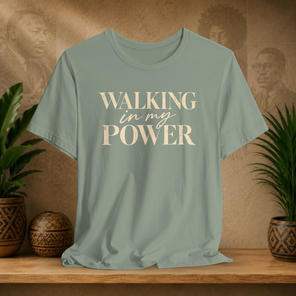 A sage green short-sleeve t-shirt with the text 'WALKING in my POWER' printed in a cream-colored serif and script font across the chest. The shirt is displayed on a wooden shelf flanked by potted green plants against a textured brown wall with sepia portraits.