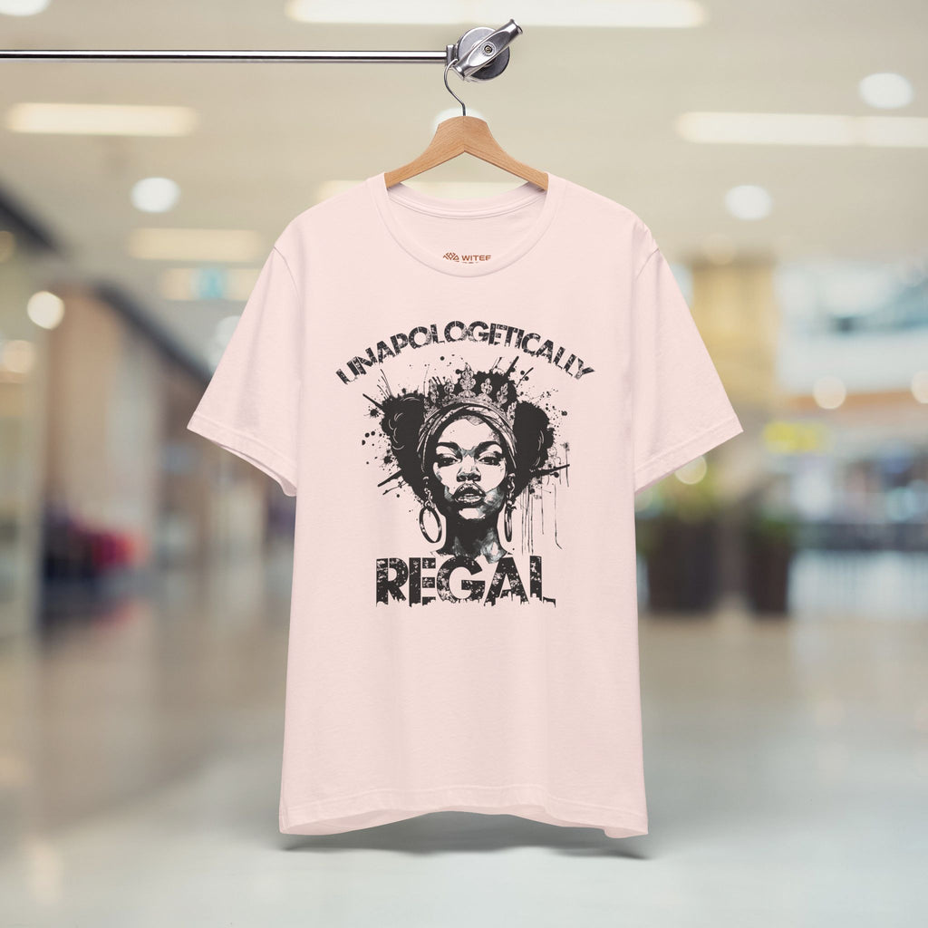 A light pink graphic t-shirt hanging on a wooden hanger from a metal rack, set against a blurred indoor shopping mall background. The t-shirt features a black distressed graphic of a woman's face with the text 'UNAPOLOGETICALLY REGAL' printed around it.