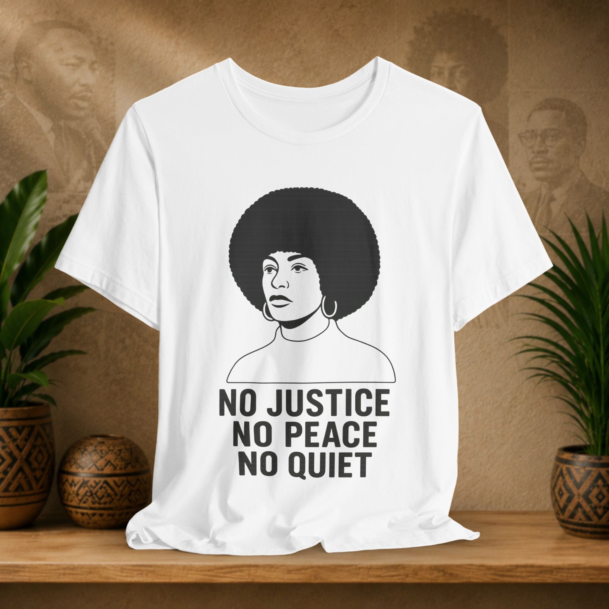 White t-shirt with a graphic of a woman with an afro and text 'No Justice No Peace No Quiet' on a wooden surface with plants and a textured wall in the background.
