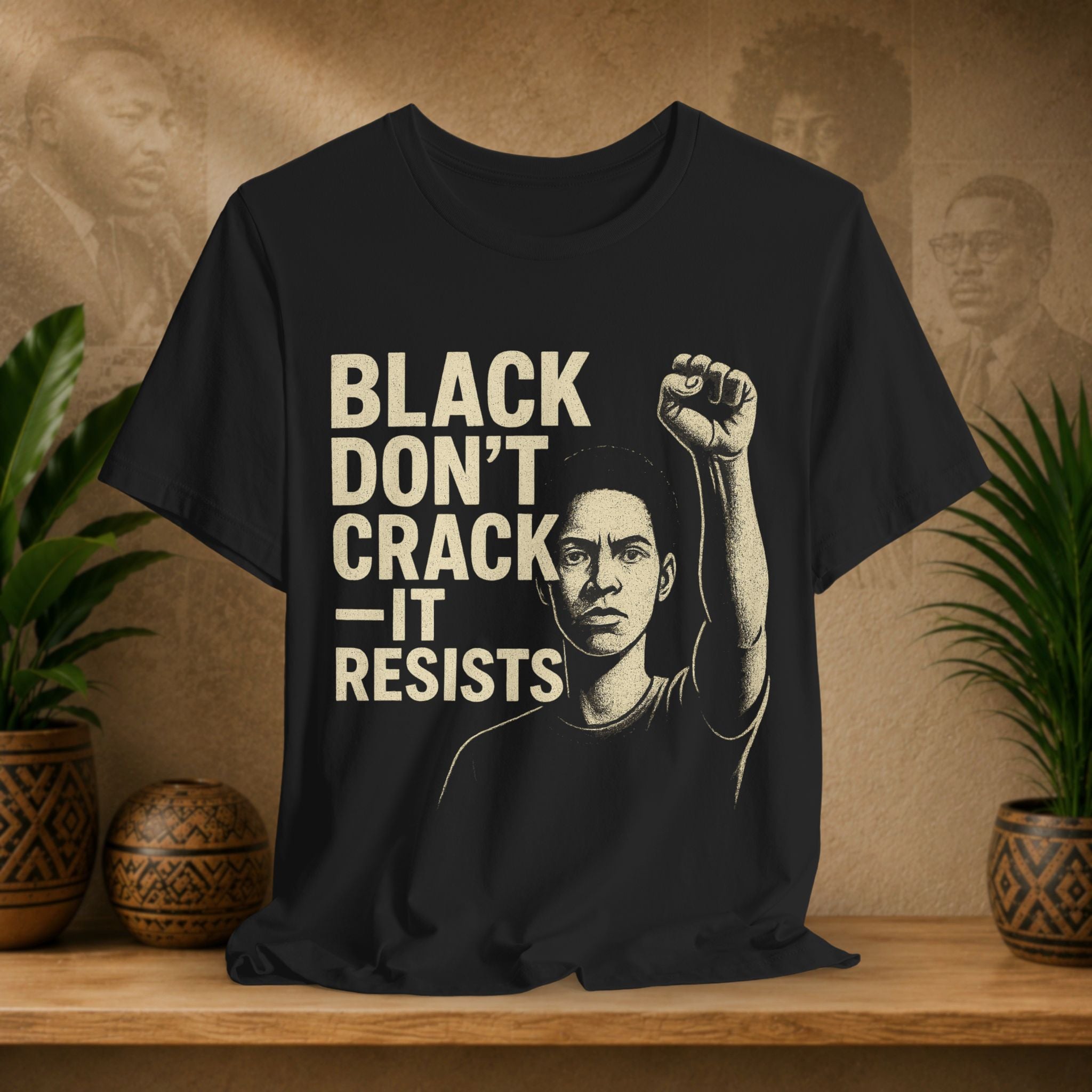 Black t-shirt with a graphic of a person raising a fist and text 'BLACK DON'T CRACK IT RESISTS' on a wooden surface with plants and decorative items.