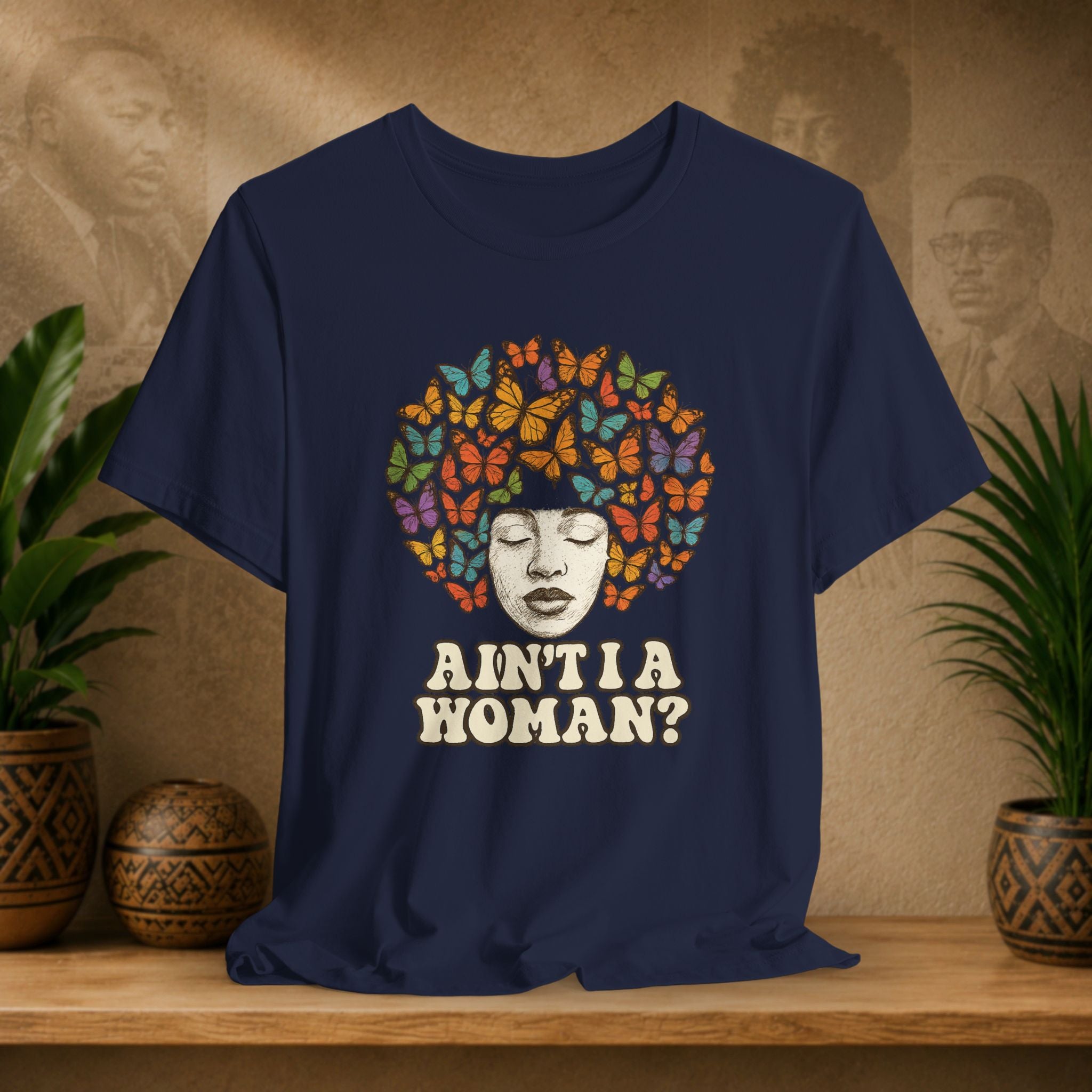 A navy blue graphic t-shirt displayed on a wooden shelf. The shirt features a graphic of a Black woman's face surrounded by a vibrant multicolor afro made of butterflies, with the text 'AIN'T I A WOMAN?' printed below in a distressed cream font.