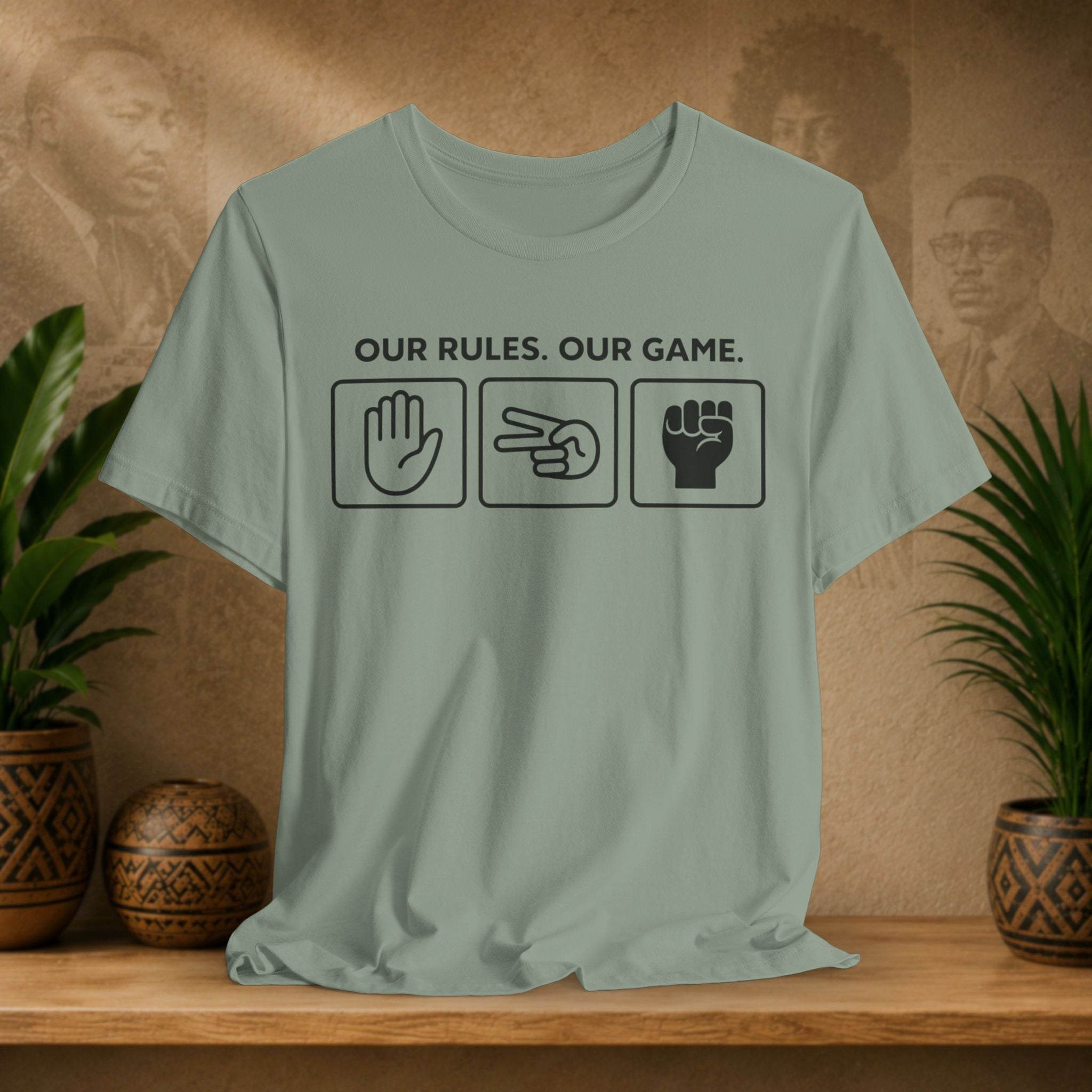 Sage green t-shirt with text and symbols on a wooden surface with plants and a textured wall background