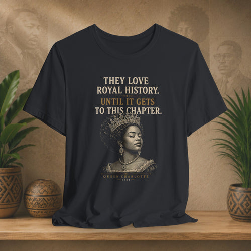 Black t-shirt with a graphic of a woman in a crown and text on a wooden surface with plants and a decorative wall.