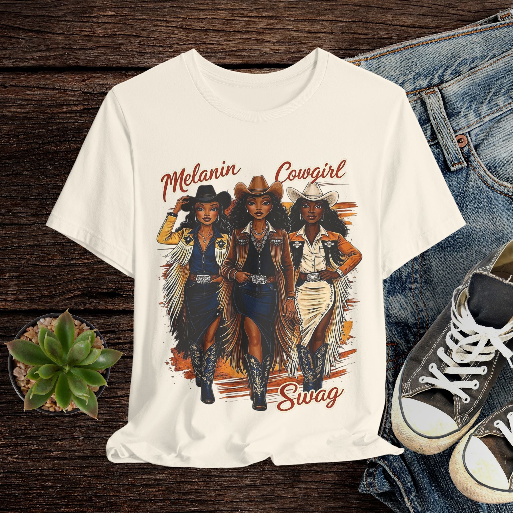 Beige t-shirt with 'Melanin Cowgirl Swag' design featuring three cowgirls on a wooden surface with jeans and sneakers.