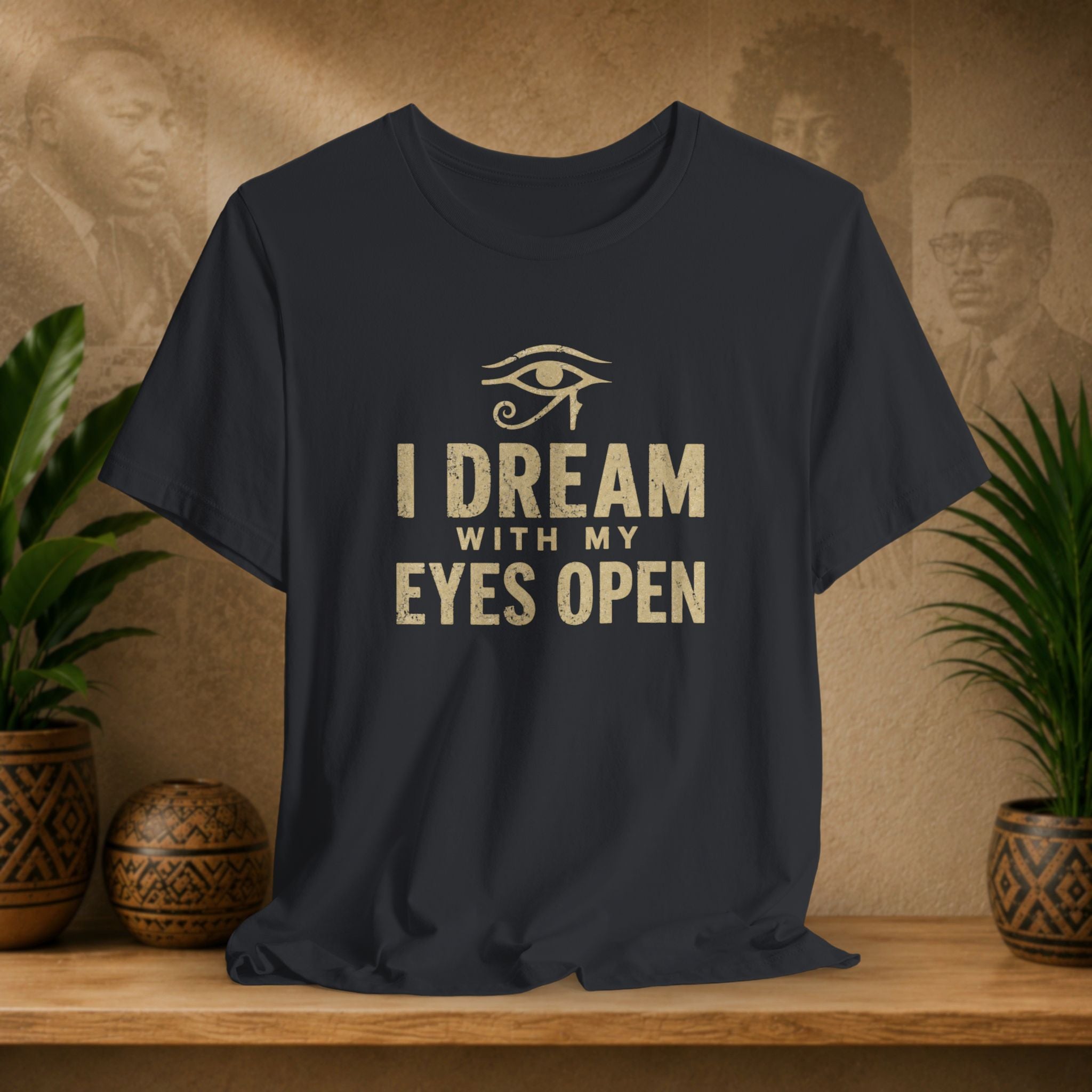 Black t-shirt with 'I Dream with My Eyes Open' text and eye symbol on a wooden surface with plants and decorative items.