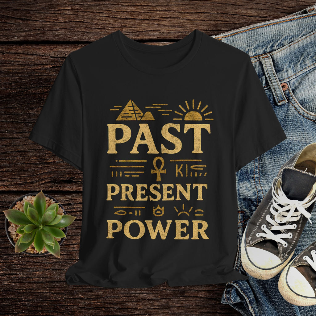 Black t-shirt with gold text 'Past is Present & Power' on a wooden surface with jeans and sneakers.
