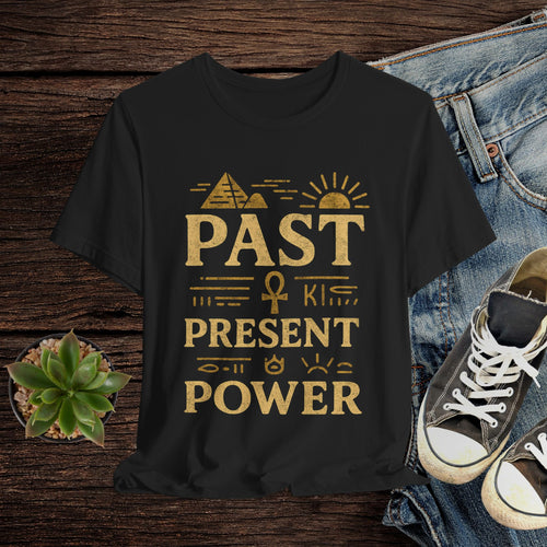 Black t-shirt with gold text 'Past is Present & Power' on a wooden surface with jeans and sneakers.