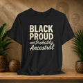 Black t-shirt with 'BLACK PROUD and Probably Ancestral' text on a wooden surface with plants and a textured wall background.