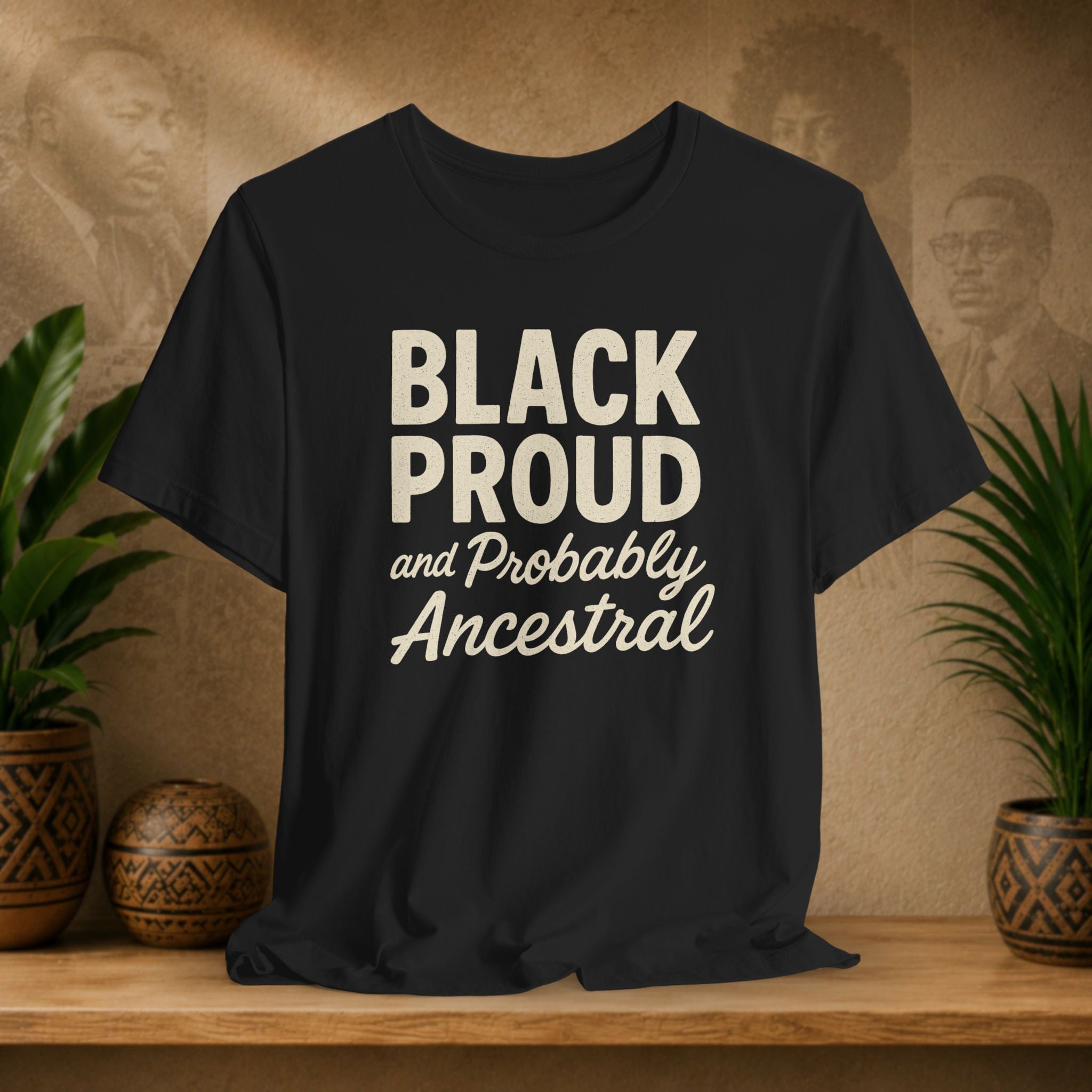 Black t-shirt with 'BLACK PROUD and Probably Ancestral' text on a wooden surface with plants and a textured wall background.