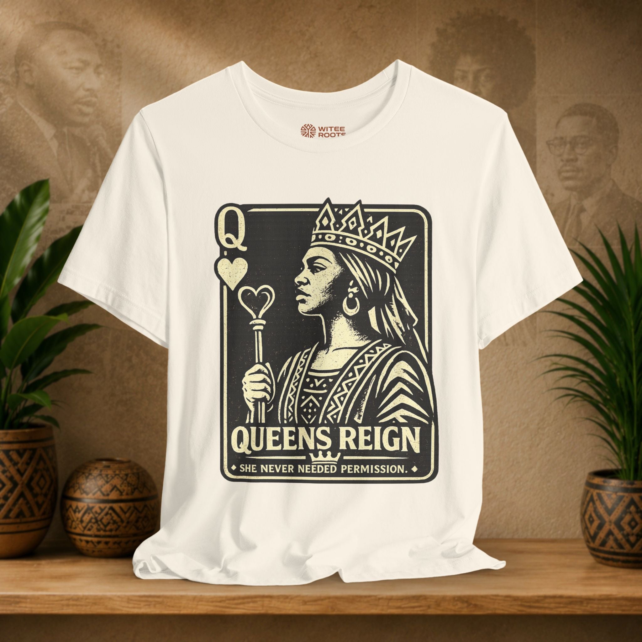 Beige t-shirt with 'Queens Reign' graphic design on a wooden surface with plants and a textured wall background.