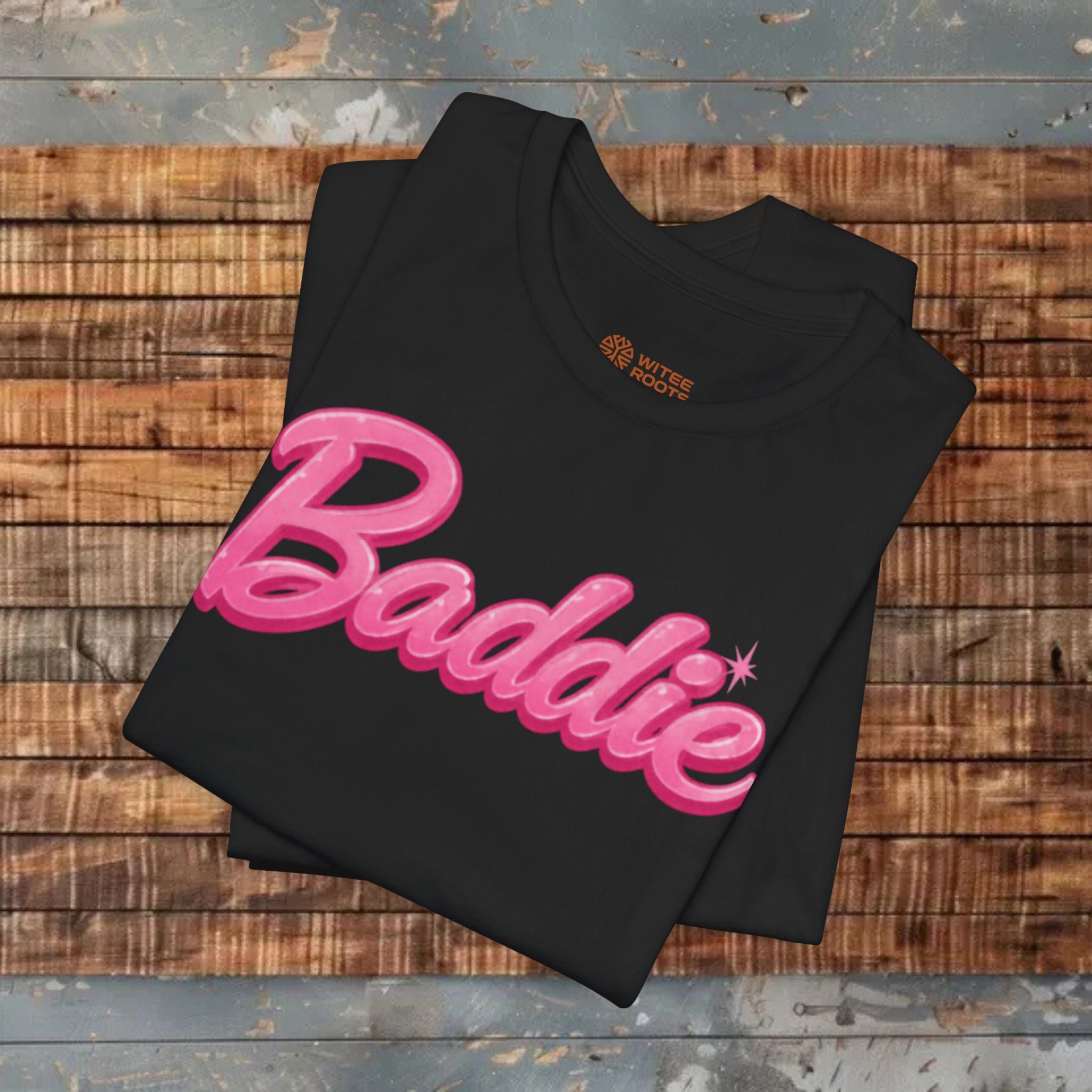 A folded black t-shirt with a pink 'Baddie' graphic printed across the front, displayed on a rustic wooden plank surface.